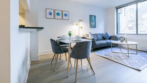 A modern living room with a dining area, a sofa, and a large window with a city view. - Fully Furnished 1BR in Murray Hills (New York)