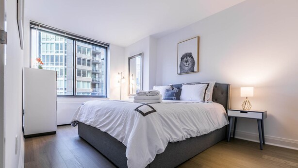 A modern bedroom with a large bed, a bedside table with a lamp, a window with a city view, and a framed picture on the wall. - Fully Furnished 1BR in Murray Hills (New York)