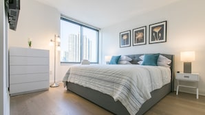 A modern bedroom with a large bed, bedside lamps, a dresser, and a window with a city view. - Bright 2 Bedrooms 2 Bathrooms Unit With Balcony (New York)