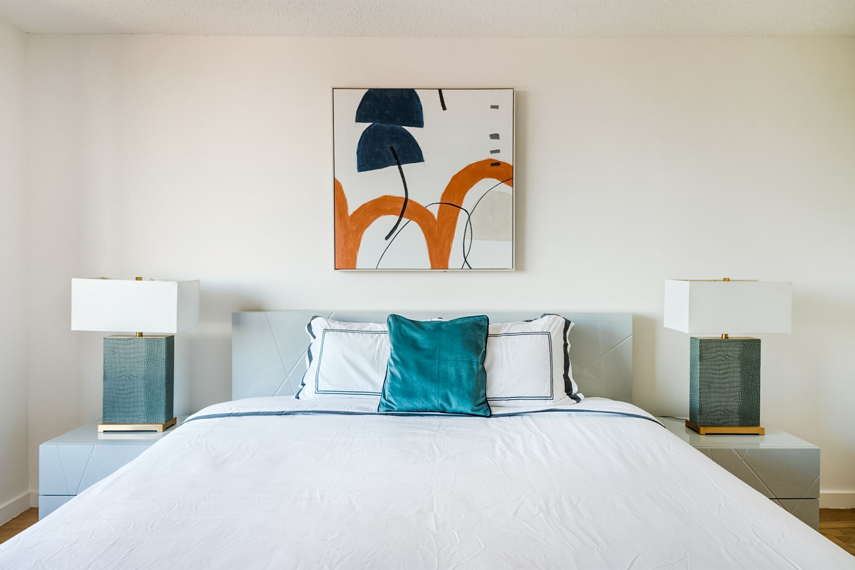 A neatly made bed with a white comforter and teal accent pillows, flanked by two bedside tables with lamps and a modern abstract painting above.