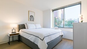 A bedroom with a bed, a nightstand, a lamp, a framed picture on the wall, and a window with a view of buildings. - Beautiful 1 Bed Fully Furnished Apartment (New York)