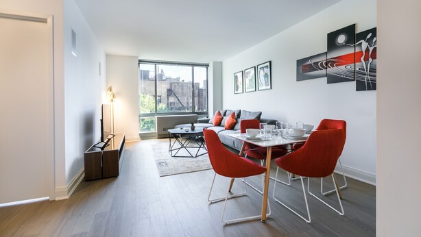 A modern dining area with a table set for four, red chairs, and a sofa in the background. - Beautiful 1 Bed Fully Furnished Apartment (New York)