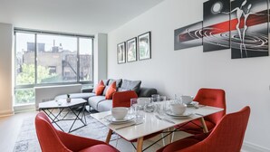A modern dining area with a table set for four, a sofa, and a large window offering a city view. - Beautiful 1 Bed Fully Furnished Apartment (New York)