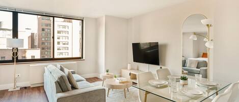A modern living room with a sofa, a glass dining table set for four, a flat-screen TV, and a large window with a city view.