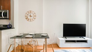 A modern kitchen with a microwave, a wall clock, and a dining area with a table, chairs, and a flat-screen TV. - Prime Upper West Side Location 1 Bedroom Apt (New York)