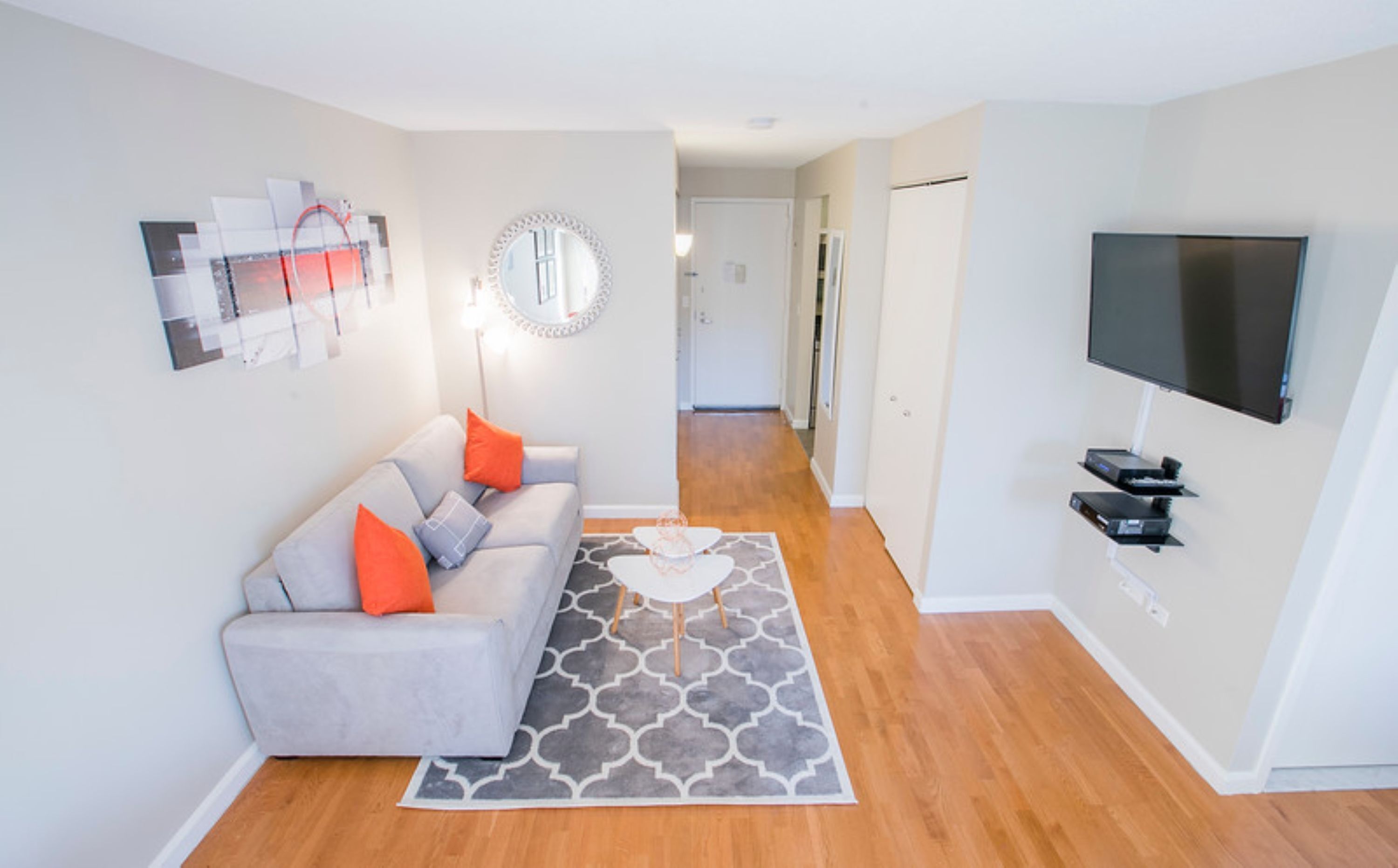 A modern living room with a grey sofa, orange cushions, a patterned rug, a flat-screen TV, and a wall-mounted mirror.