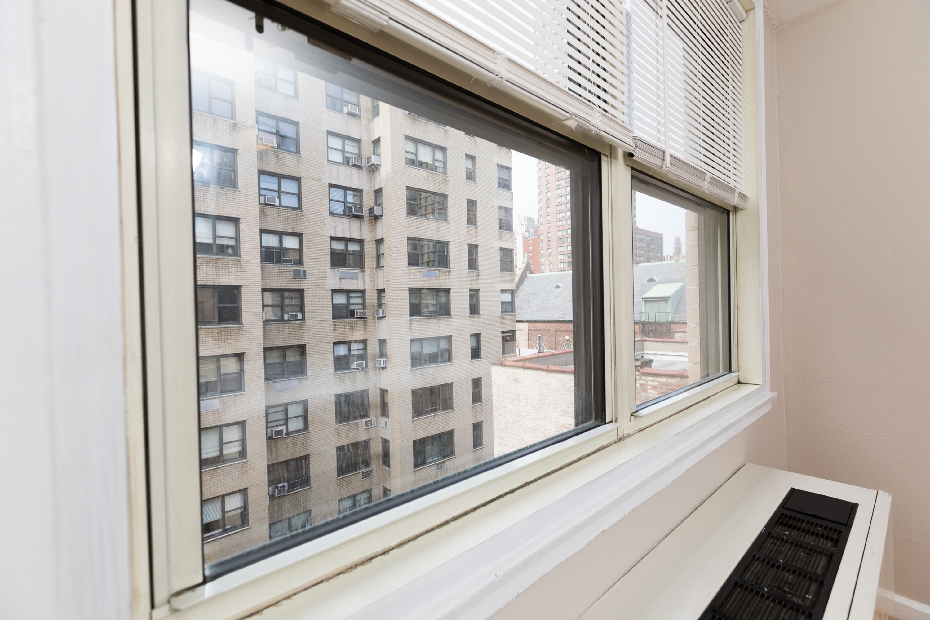 Ett fönster med utsikt över en hög byggnad, ett ventilationsgaller och en fönsterkarm.