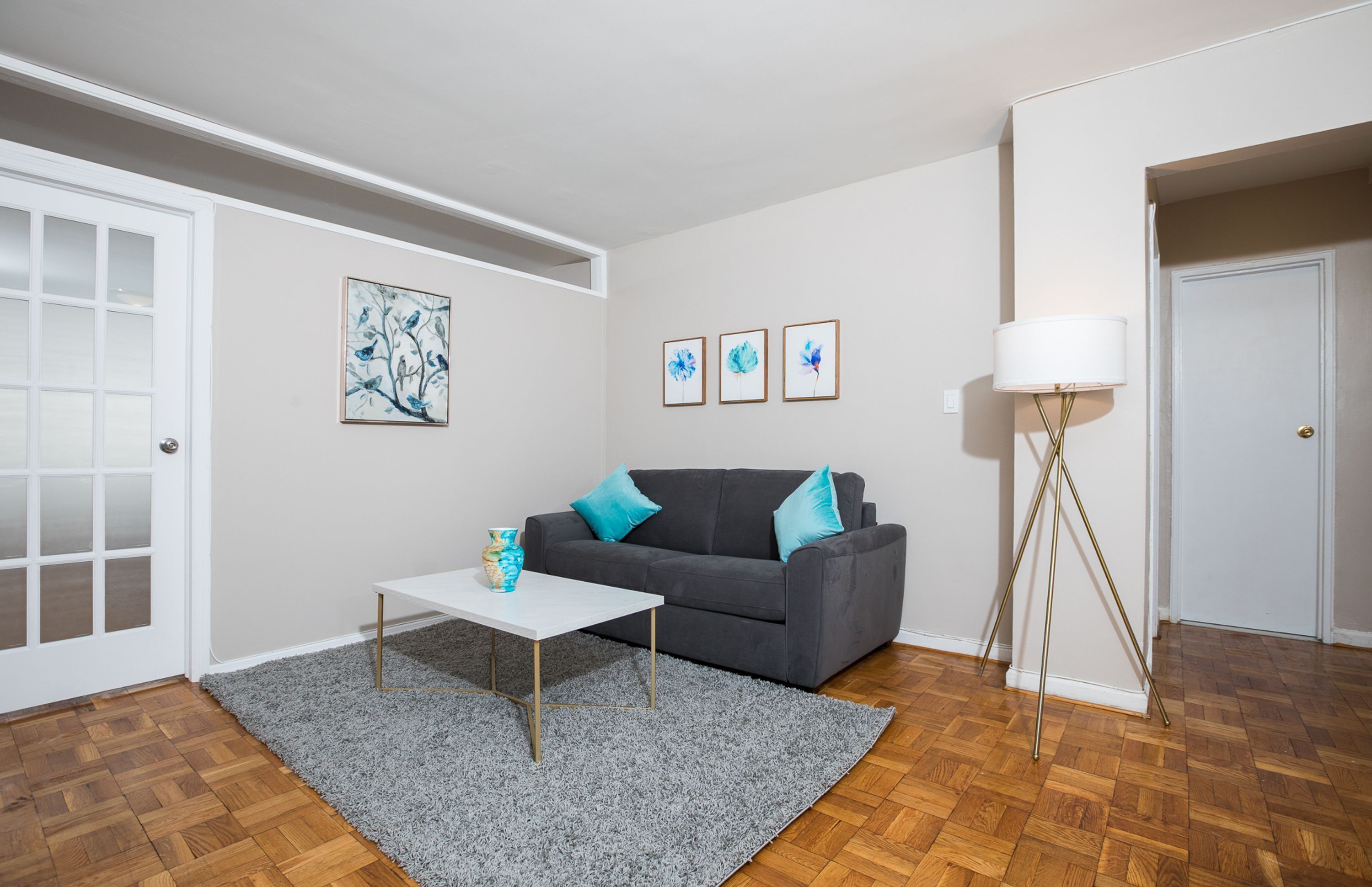 A living room with a grey sofa, a white coffee table, and a grey rug. There are three framed pictures on the wall and a floor lamp.