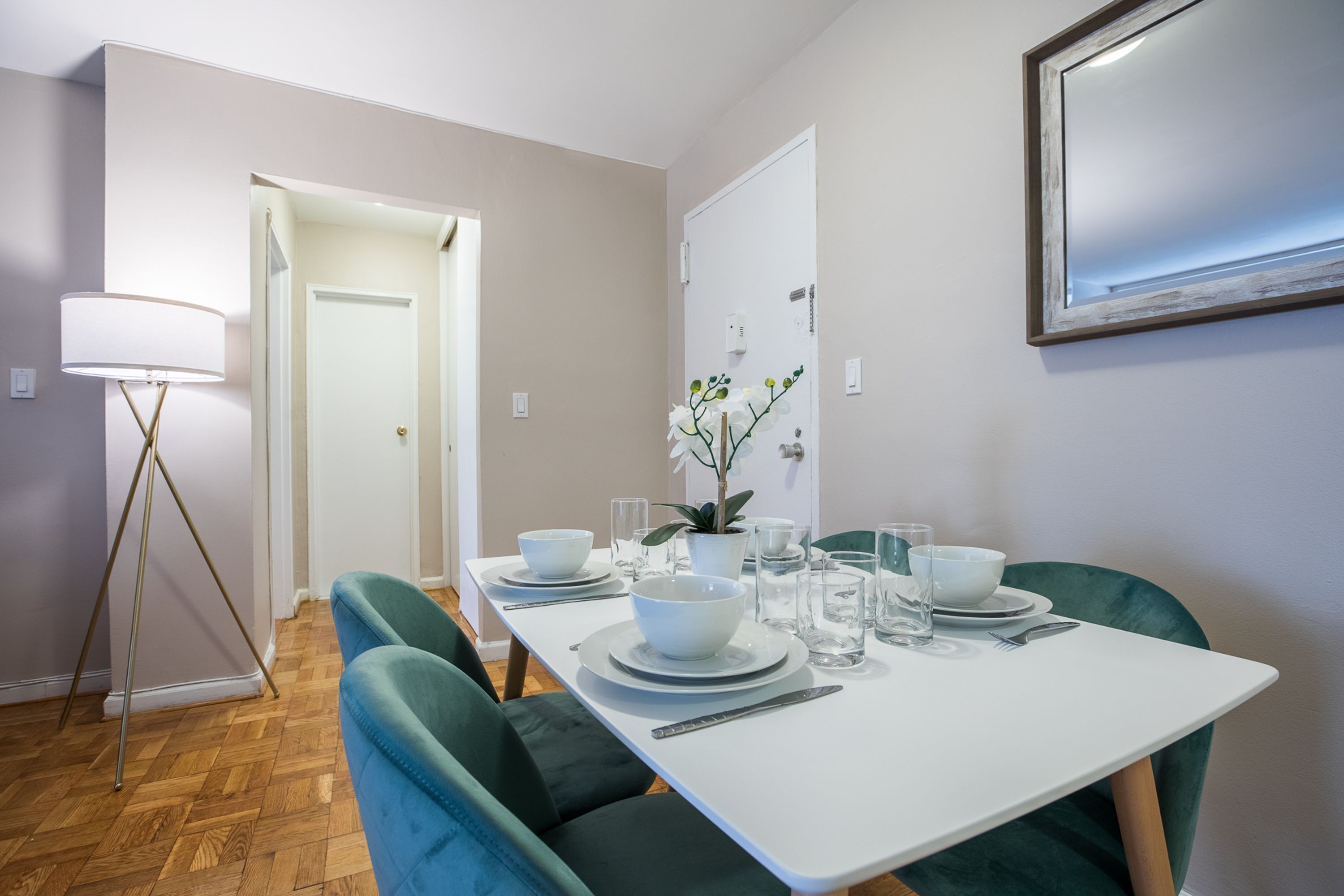 Uma sala de jantar com uma mesa branca posta para quatro pessoas, cadeiras estofadas verdes, uma orquídea em vaso e um abajur.