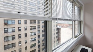 A view through a window with blinds, revealing a multi-story building with numerous windows and air conditioning units. - 2 Bed Apt Ideally Located in UES of Manhattan (New York)