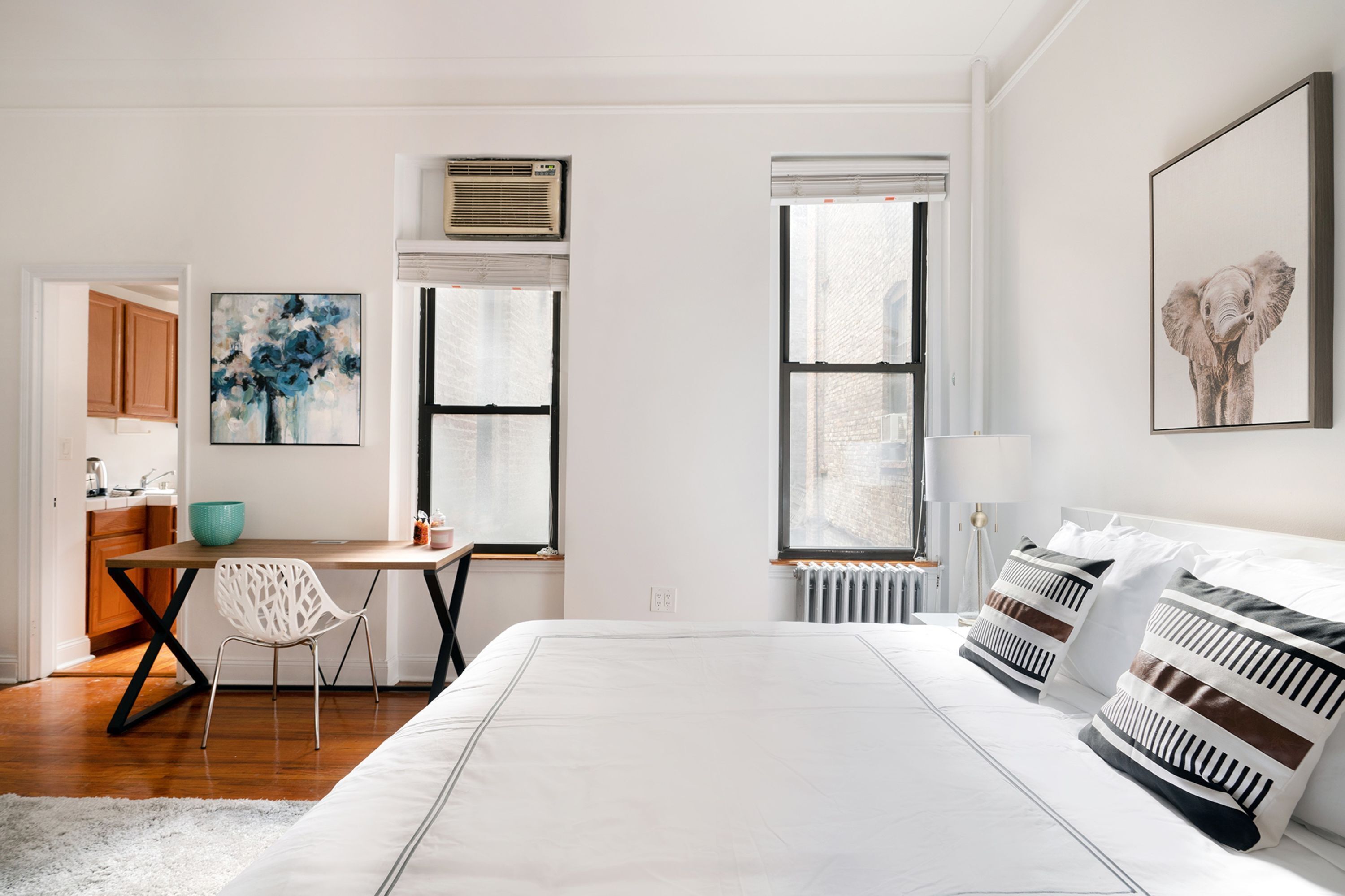 Un dormitorio moderno con una cama grande, un escritorio y una silla. Hay dos ventanas, una con persianas y otra con vistas a la ciudad.