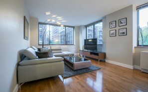 Una sala de estar moderna con un sofá de cuero, una mesa de centro y un televisor de pantalla plana sobre un mueble de madera. Amplias ventanas ofrecen vistas de la ciudad.