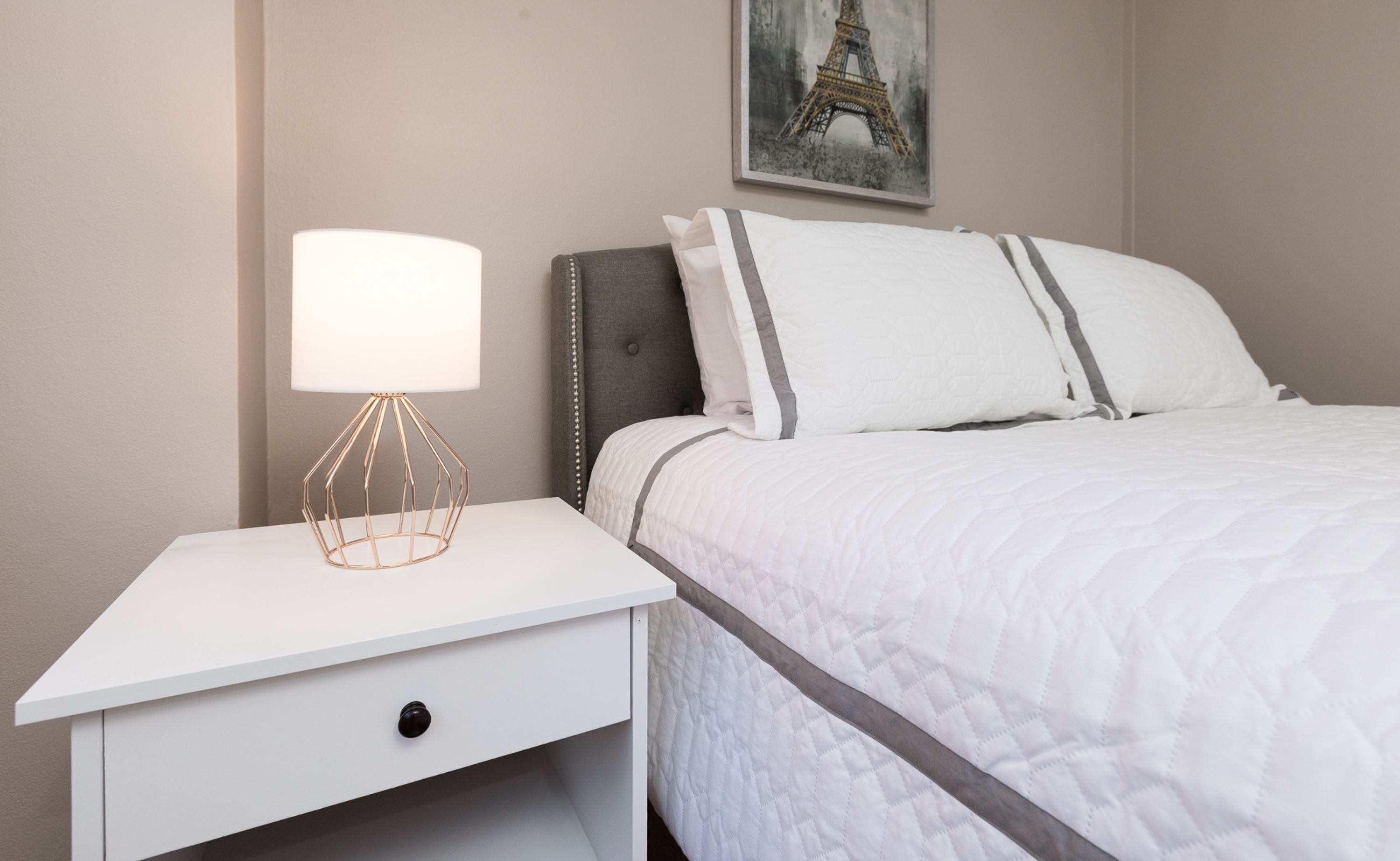 A neatly made bed with white bedding and gray stripes, a white nightstand with a gold lamp, and a painting of the Eiffel Tower on the wall.