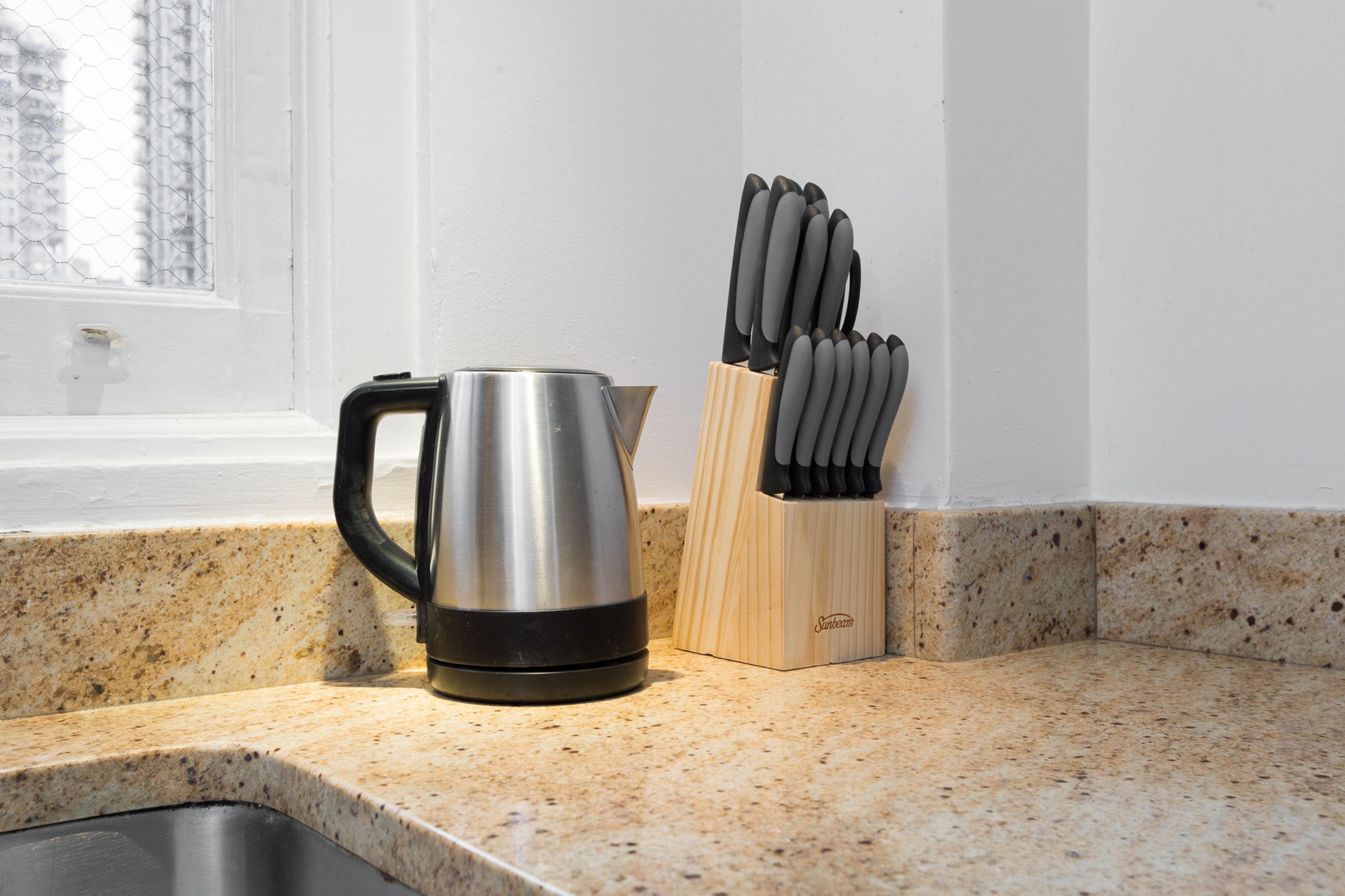 A kitchen counter with a stainless steel kettle, a knife block, and a window in the background.