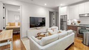 En moderne stue med sofa, sofabord og fjernsyn. Ved siden af er et køkken med køleskab, komfur og skabe.