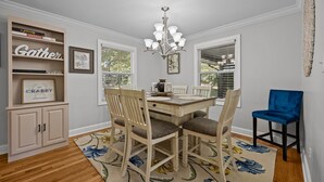 A dining area with a wooden table, chairs, and a chandelier. There is a bookshelf with books and a sign. - The Retreat at Kincaid (Raleigh)