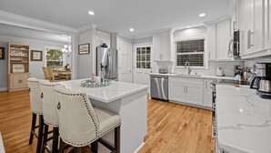 A modern kitchen with white cabinetry, stainless steel appliances, and a central island with bar stools. - The Retreat at Kincaid (Raleigh)