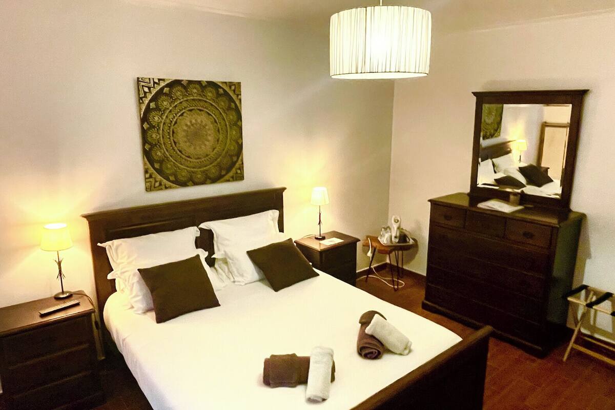 A neatly made bed with pillows, a bedside lamp, a dresser, a mirror, and a decorative wall piece.
