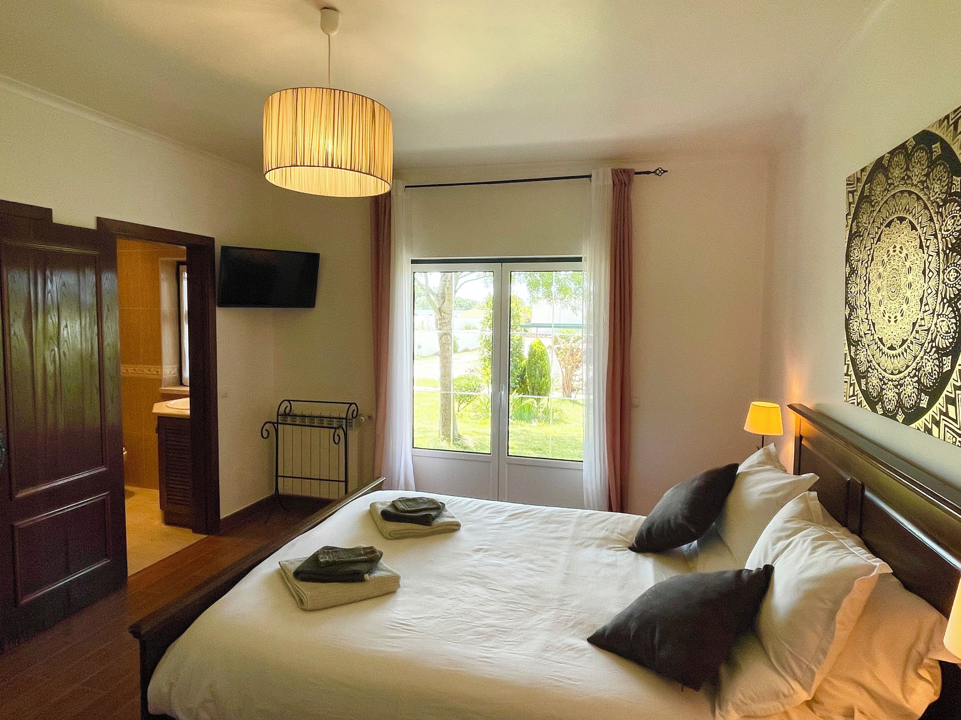 A bedroom with a bed, a bedside lamp, a window with curtains, a mounted TV, and a decorative wall piece.