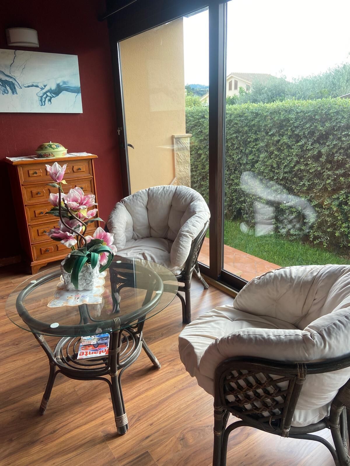A room with two wicker chairs, a glass table with a vase of flowers, a wooden dresser, and a large window with a view of greenery.