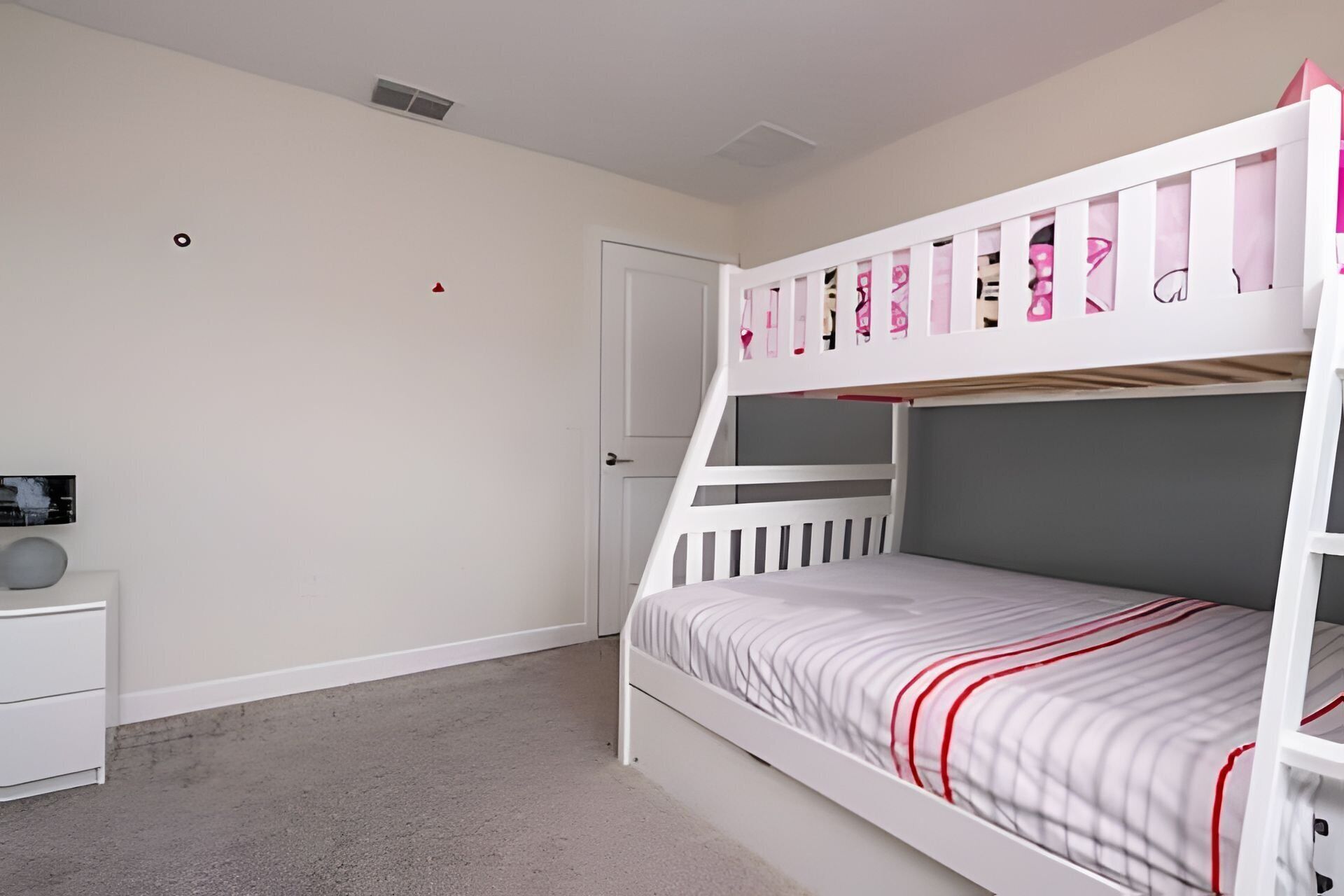 A bunk bed with a white frame and a grey mattress, a white nightstand with a round object, and a white door.