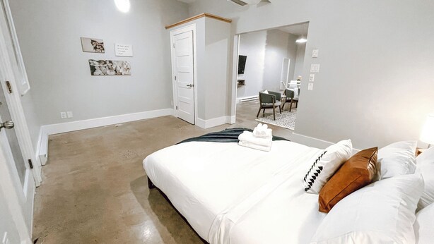 Quarto moderno com uma cama grande, uma porta para um banheiro e uma pequena área de estar com uma cadeira e mesa.