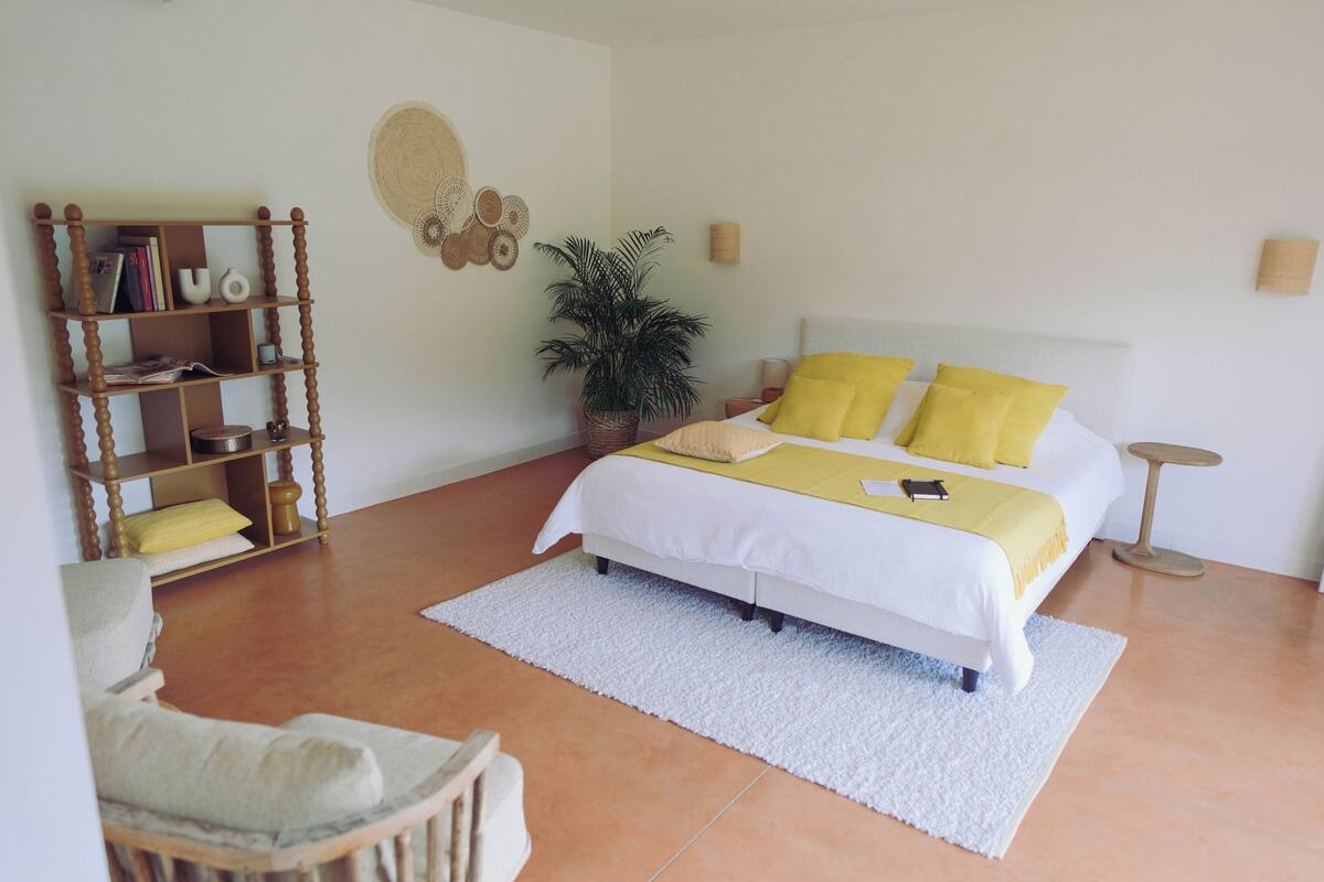 Un dormitorio con cama, mesita de noche, lámpara, estantería y una planta en maceta.