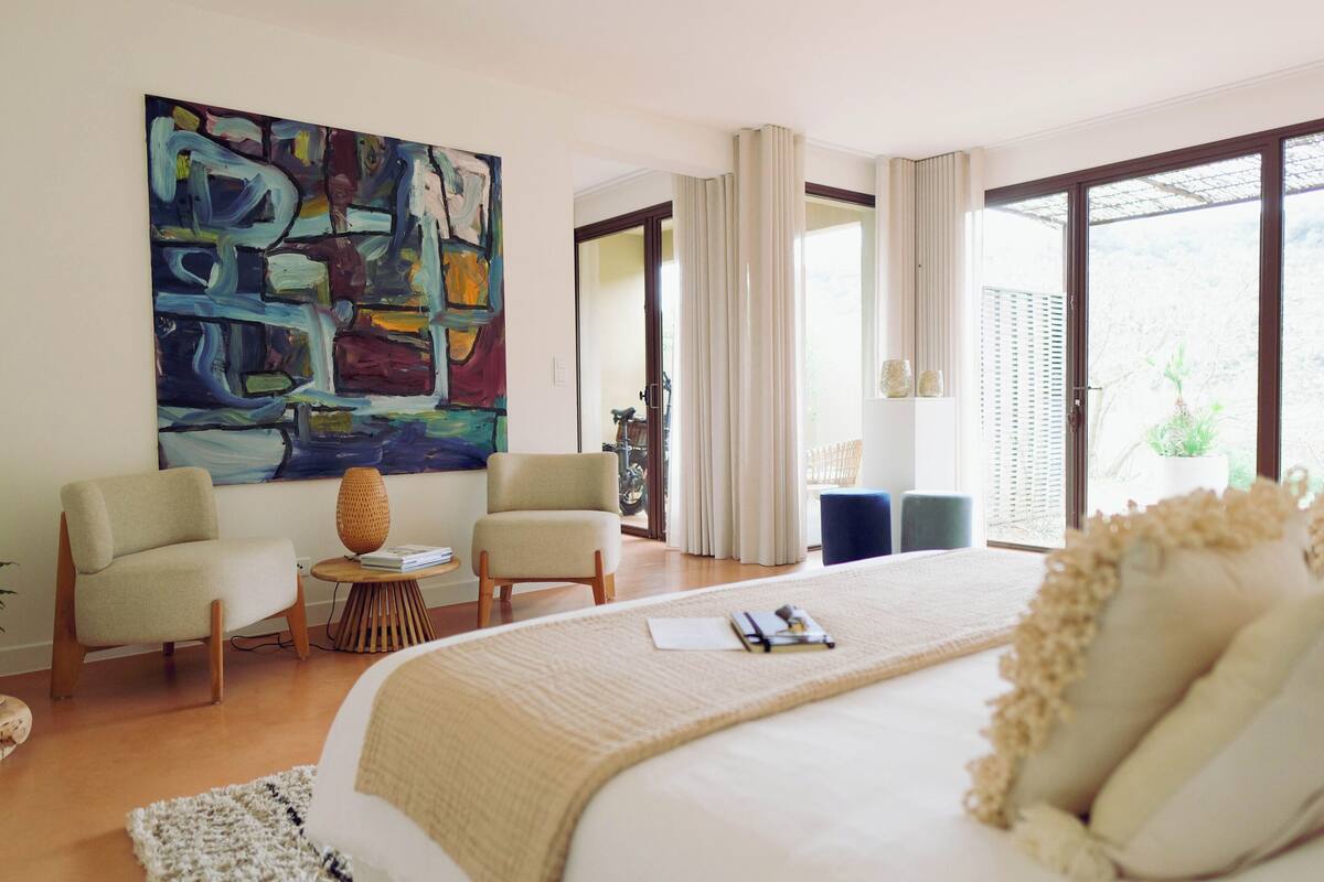 Une chambre d’hôtel moderne, dotée d’un grand lit, de deux fauteuils, d’une petite table et d’un grand tableau abstrait accroché au mur.