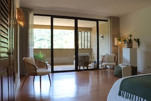 A hotel room with a large sliding glass door leading to a balcony, a bed, a chair, a small table, and a potted plant. - Louis Henry (Saint-Pierre-des-Champs)