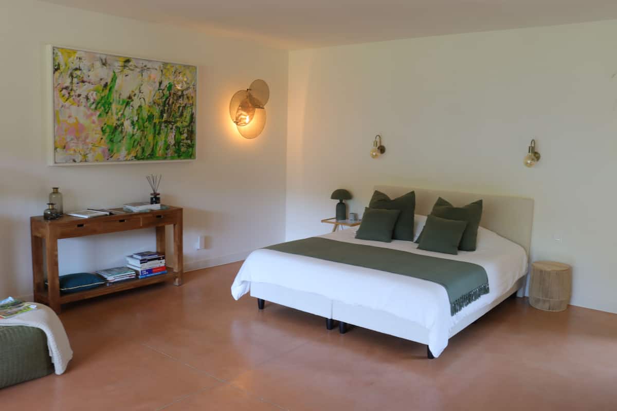 Un dormitorio con cama, mesita de noche, luz en la pared, un cuadro y un taburete de madera.