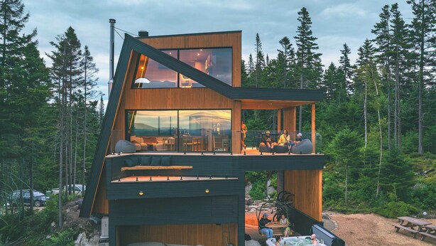 A modern multi-level cabin with a large glass window, outdoor seating, and a hot tub.