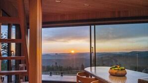 A modern kitchen with a large window overlooking a scenic view, wooden chairs, and a fruit bowl on the counter.