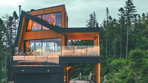A modern two-story cabin with a glass balcony, outdoor seating area, and a hot tub.