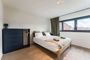 A modern bedroom with a bed, a dark wardrobe, a window with curtains, and a view of buildings outside. - Cosy & Spacious House in Center (Kortrijk)