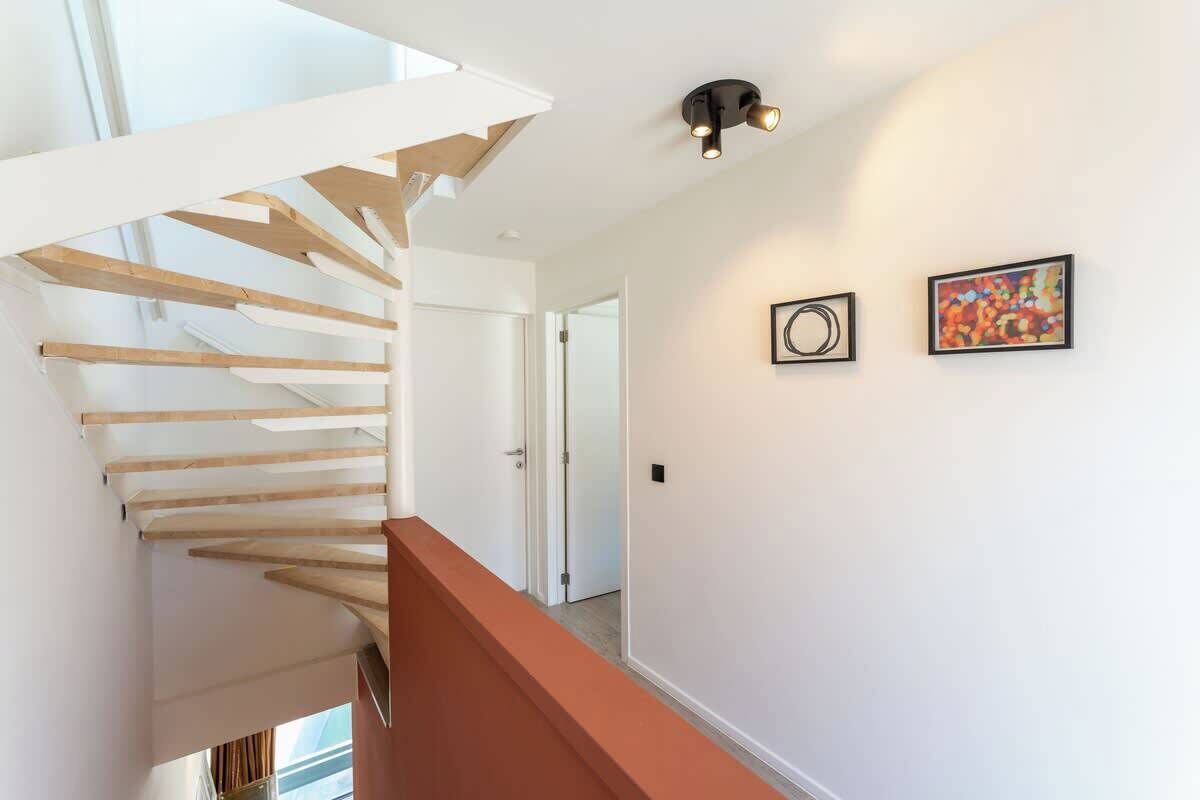 Un escalier moderne avec des marches en bois et une rampe rouge, menant à une pièce aux murs blancs et ornée de deux œuvres d’art encadrées.