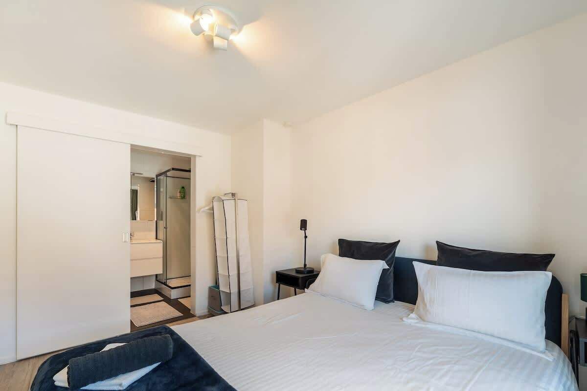 Une chambre d’hôtel avec un grand lit, une table de chevet avec une lampe, une armoire et une salle de bain visible à travers une porte ouverte.