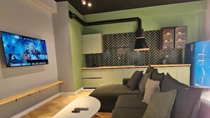 Ein modernes Wohnzimmer mit großem Flachbildfernseher, einem grauen Sofa, einem weißen Couchtisch und einer Küchenzeile mit Zickzack-Bodenfliesen und Schränken.