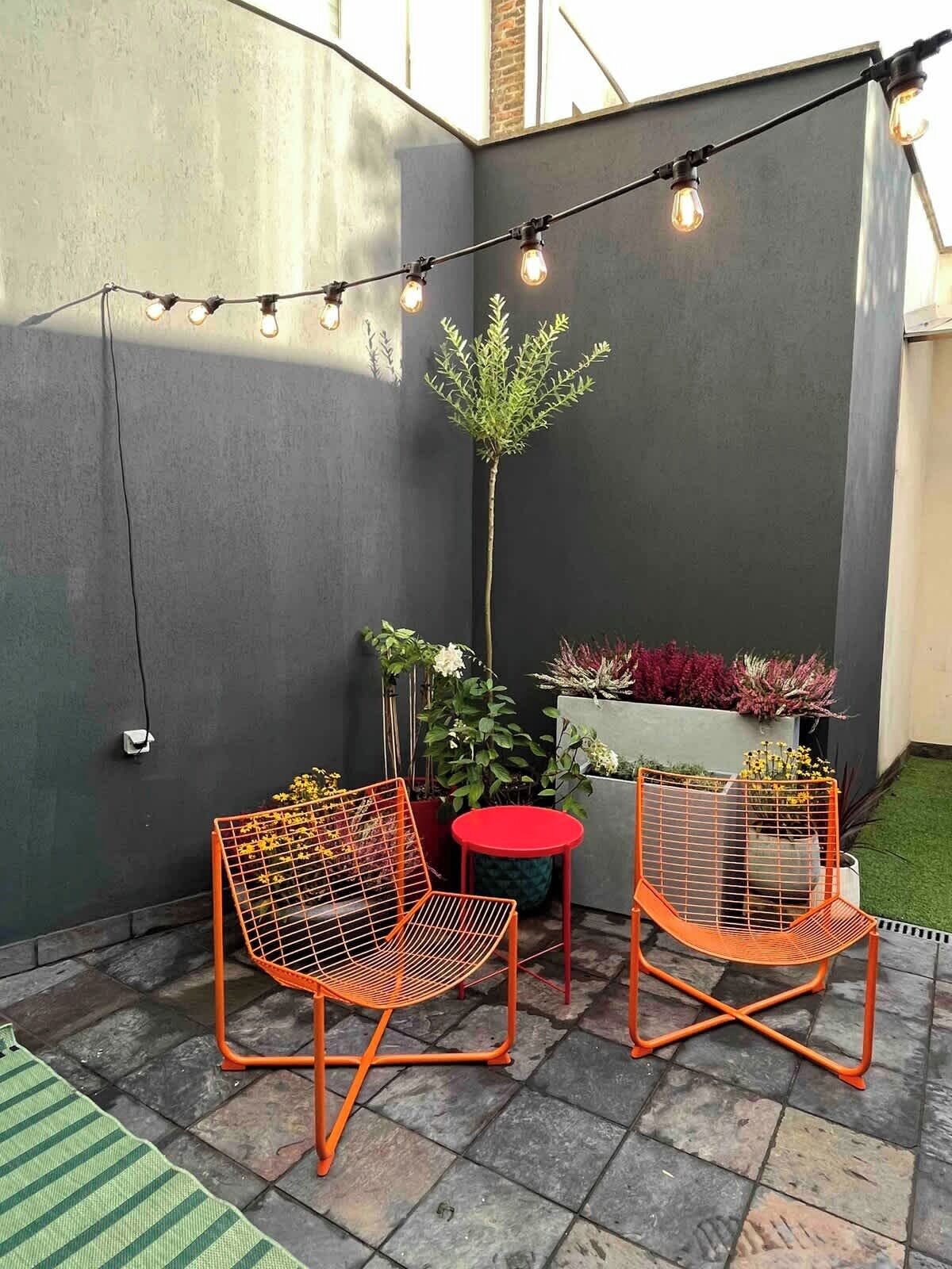 An outdoor seating area with two orange wire mesh chairs, a small red table, and a string of lights.