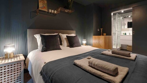 Une chambre à coucher moderne avec un grand lit, des lampes de chevet, une table de chevet, un miroir et une petite étagère ornée d’objets décoratifs.