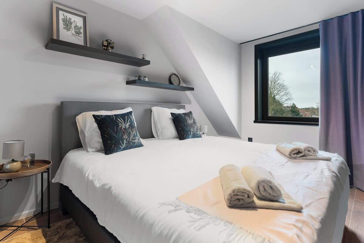 Un dormitorio moderno con una cama grande, dos almohadas, una mesita de noche con una lámpara y una ventana con cortinas.