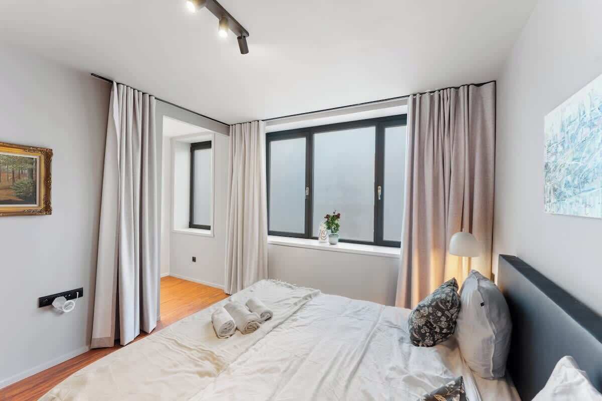 A modern bedroom with a large bed, a window with curtains, a bedside table with a lamp, and a painting on the wall.