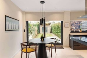 Un espace repas moderne avec une table ronde, deux chaises, un luminaire suspendu et une vue sur un jardin grâce à de grandes portes vitrées.