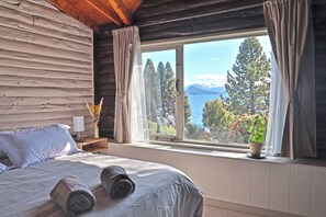 A wooden cabin room with a bed, a nightstand, a window with curtains, and a view of trees and mountains. - Casa VanTitter (San Carlos de Bariloche)