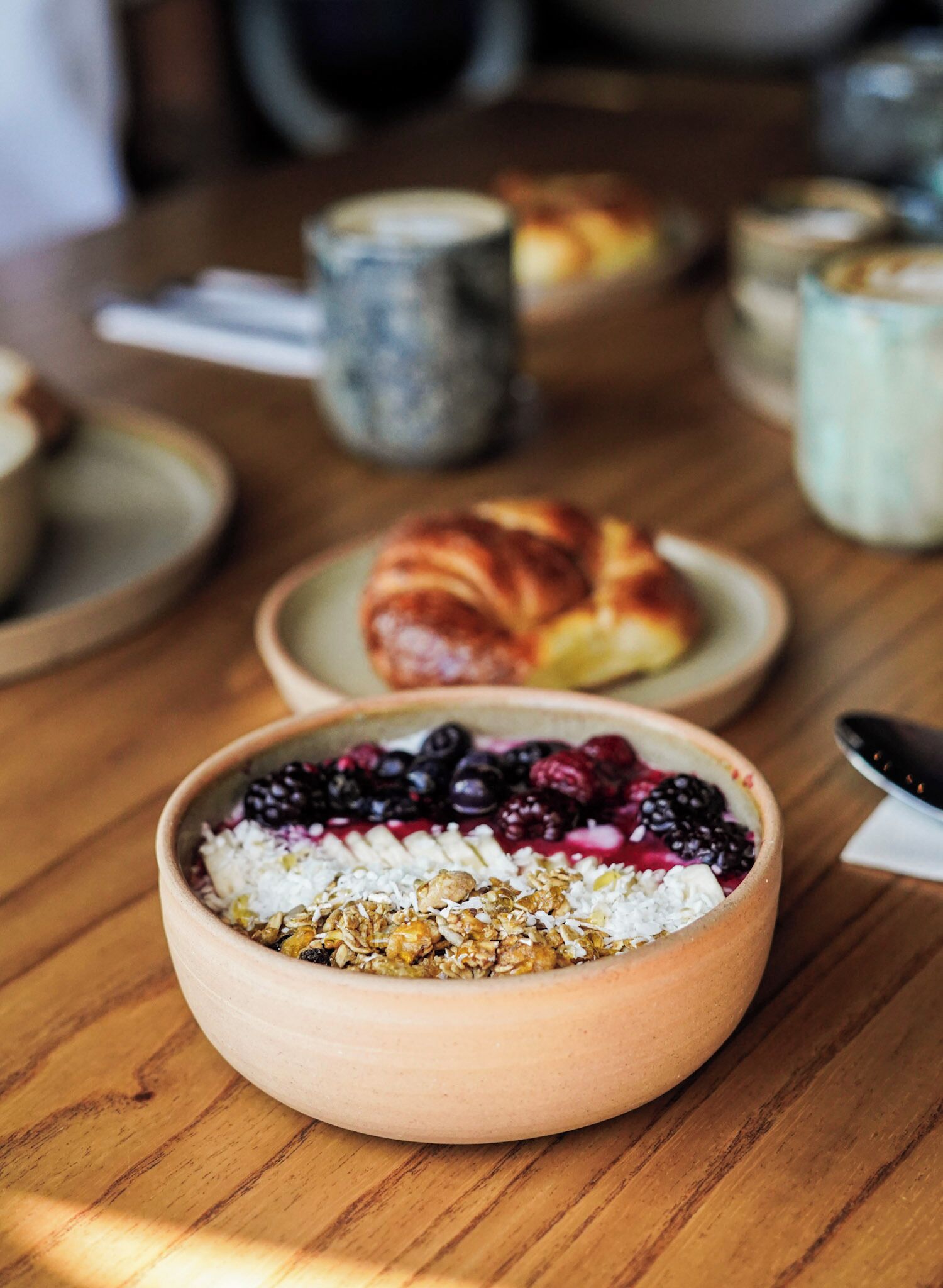 A wooden table with a bowl of cereal topped with fruits and nuts, a croissant, and a mug.