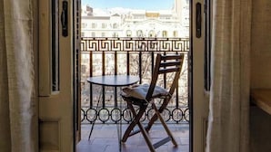 Een balkon met een tafel en stoel, een smeedijzeren hekwerk, en een uitzicht op gebouwen op de achtergrond.