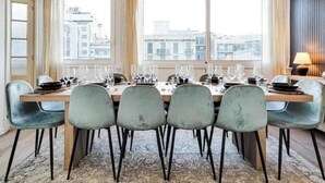 Een eetruimte met een lange tafel voor zes personen, grote ramen met gordijnen en uitzicht op gebouwen.