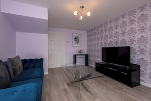 A living room with a blue sofa, a glass coffee table, a black entertainment unit, and a television. - Toast Stays - Durham View (Spennymoor)