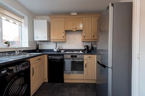 A modern kitchen with wooden cabinets, a stainless steel refrigerator, a dishwasher, and a built-in oven. - Toast Stays - Durham View (Spennymoor)