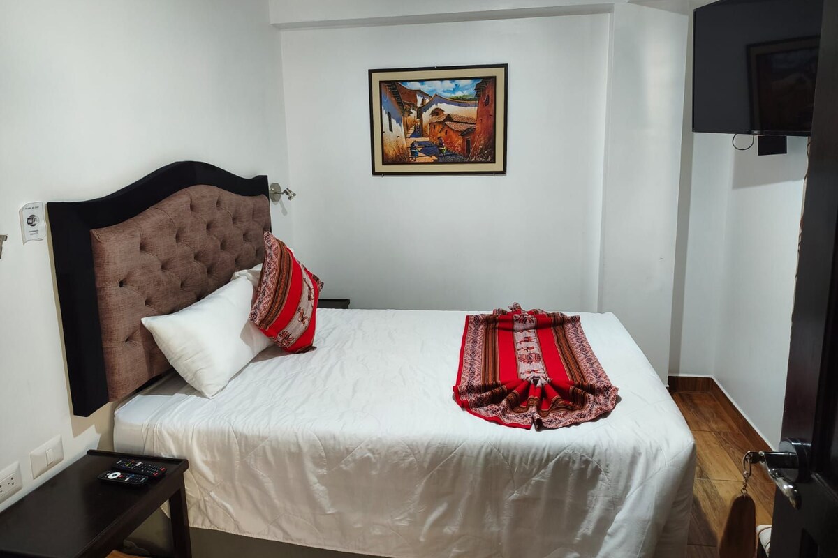 Un dormitorio con una cama individual, cabecera, mesita de noche, control remoto, un cuadro enmarcado en la pared y una puerta con picaporte.