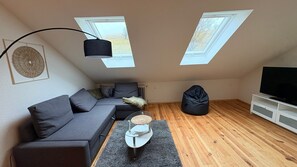 Ein Loft-Zimmer mit grauem Ecksofa, einem Glastisch, einem schwarzen Sitzsack, einer weißen TV-Konsole und Dachfenstern.